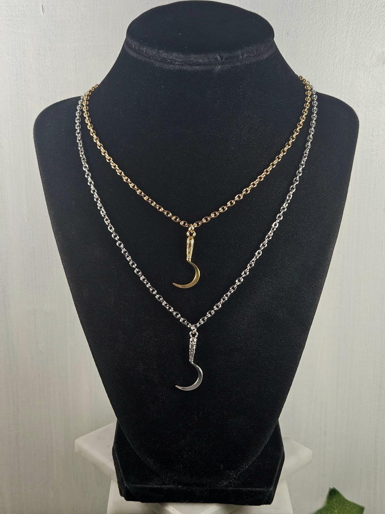 May include: Two necklaces on a black display. One necklace is gold-toned, the other is silver-toned. Each necklace features a crescent moon pendant. The chains are delicate and the pendants are small, creating an elegant look.