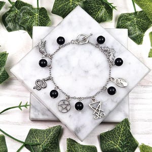 May include: A silver charm bracelet with black beads and various charms, including a snake, a pentagram, a crescent moon, and a winged creature. The bracelet is on a white marble surface.