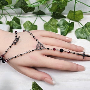 May include: A black and red beaded finger ring with a Celtic knot charm and a sword charm. The ring is made of metal and has a delicate chain.