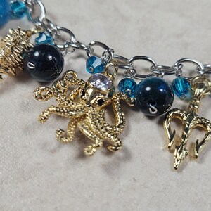 May include: A silver chain bracelet with various charms. Charms include a gold seahorse, a gold octopus with a clear gem, and a gold trident. The bracelet also features blue and dark blue beads.