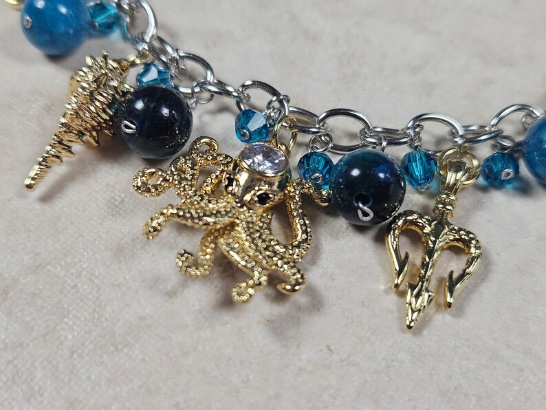 May include: A silver chain bracelet with various charms. Charms include a gold seahorse, a gold octopus with a clear gem, and a gold trident. The bracelet also features blue and dark blue beads.