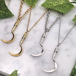 May include: Four necklaces with a sickle pendant. Two are gold-colored, and two are silver-colored. Each pendant is attached to a chain. The necklaces are displayed on a white marble surface, with green leaves in the background.
