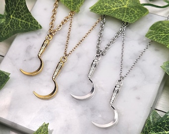 Puede incluir: Cuatro collares con colgantes de hoz. Dos son dorados y dos plateados. Cada colgante está unido a una cadena. Los collares se exhiben sobre una superficie de mármol blanco, con hojas verdes en el fondo.