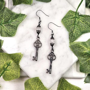 Puede incluir: Un par de pendientes de estilo antiguo con colgantes de llave. Los pendientes tienen un acabado de metal oscuro, una cuenta transparente y un dije de llave decorativa. Se exhiben sobre una superficie de mármol blanco con hojas verdes.