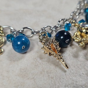 May include: A silver chain bracelet with ocean-themed charms. The charms include gold seahorses, a seashell, and a starfish. Blue beads and crystals are interspersed throughout the chain, creating a nautical aesthetic.