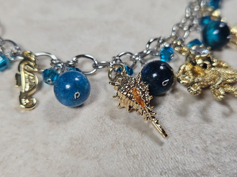 May include: A silver chain bracelet with ocean-themed charms. The charms include gold seahorses, a seashell, and a starfish. Blue beads and crystals are interspersed throughout the chain, creating a nautical aesthetic.