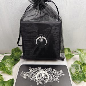 Op de afbeelding: Een zwarte juwelendoos met een halve maan ontwerp op de voorkant, verpakt in een zwarte gaas zak. De doos staat op een zwarte kaart met een witte halve maan en een bloemmotief, en de tekst "Jennifer Nicole Jewelry".