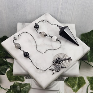 May include: Black pendulum with a black and white crystal chain. The chain has a key, a labyrinth, and a crescent moon charm. The pendulum is on a white surface.
