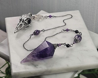 Puede incluir: Un péndulo con una punta de cristal de amatista morada facetada unida a una cadena de metal oscuro. La cadena incluye pequeñas cuentas negras y moradas, un dije de pentáculo plateado y un colgante de cráneo de ave plateado en el extremo.