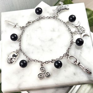 Peut inclure: Un bracelet à breloques en argent avec des perles noires et divers breloques, dont un crâne, une épée, un symbole de triple lune, un triskèle, une femme tenant une baguette et un oiseau.