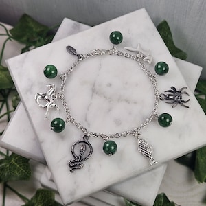 Könnte beinhalten: Ein silbernes Charm-Armband mit dunkelgrünen Perlen und verschiedenen Anhängern, darunter eine Feder, ein Seepferdchen, ein Fisch, eine Spinne und eine Schlange. Das Armband wird auf einer weißen Marmoroberfläche präsentiert.