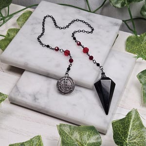 May include: Black pendulum with a silver charm featuring a symbol. The pendulum is attached to a black chain with red beads.