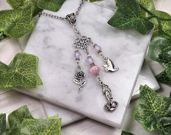 May include: A silver-toned necklace with a floral design, pink beads, and charms of a rose, bird, and mermaid. The necklace is displayed on a white marble surface, surrounded by green leaves.
