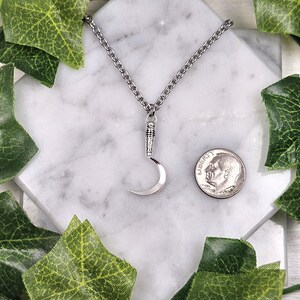 May include: Silver-toned crescent moon pendant necklace. The pendant is suspended from a chain and is displayed on a white marble surface. A U.S. dime is next to the pendant for size comparison. Green leaves surround the necklace.