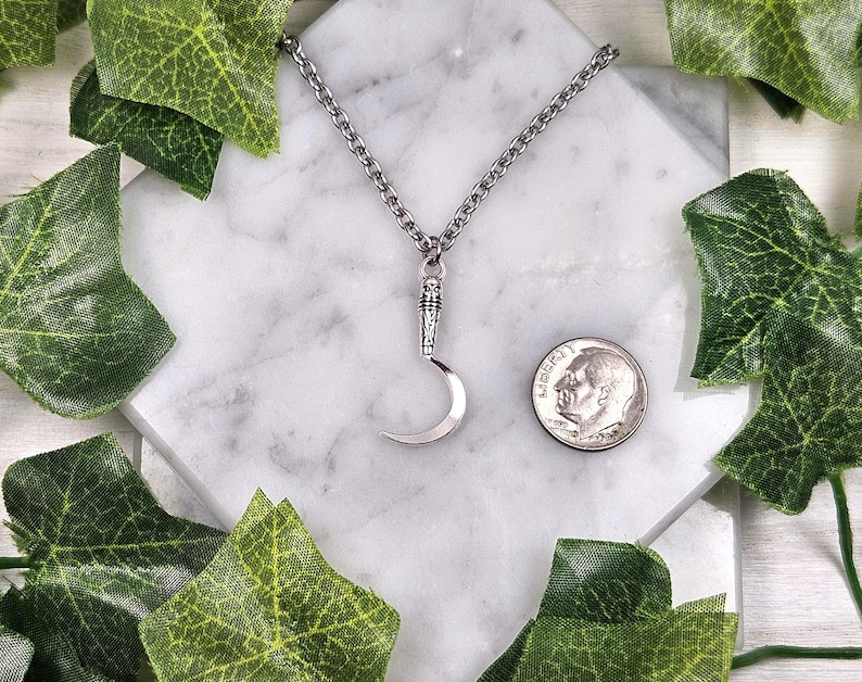 May include: Silver-toned crescent moon pendant necklace. The pendant is suspended from a chain and is displayed on a white marble surface. A U.S. dime is next to the pendant for size comparison. Green leaves surround the necklace.