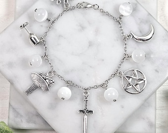 May include: Silver-toned charm bracelet featuring various charms, including a crown, a champagne flute, a mushroom, a sword, a crescent moon, and a pentagram. The bracelet also has white, round beads. The bracelet is displayed on a white marble surface.
