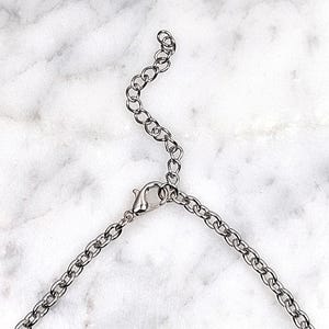 May include: Silver-toned necklace chain with a lobster clasp and adjustable extender. The chain is composed of small, oval links. The background is a white and gray marble pattern.
