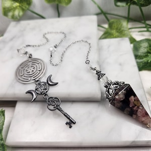 Könnte beinhalten: Ein silbernes Pendel mit einer Kristallspitze und einer silbernen Kette. Das Pendel hat einen silbernen Anhänger mit einem Schlüssel und einem Dreifachmond-Symbol. Die Kette hat einen silbernen Anhänger mit einem Labyrinth-Design.