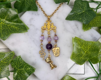 May include: Gold-toned pendant necklace featuring a decorative design with amethyst beads and charms. The charms include a snake, a theater mask, and a chalice. The necklace is displayed on a white marble surface with green leaves.