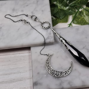 Puede incluir: Un péndulo de cristal negro con una cadena de plata y un dije de media luna. El péndulo cuelga de una cadena de plata con una cuenta decorativa en la parte superior. El dije de media luna está unido a la cadena debajo del péndulo.