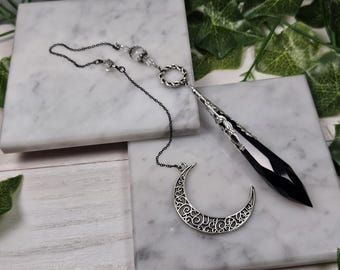 Puede incluir: Un péndulo de cristal negro con una cadena de plata y un dije de media luna. El péndulo cuelga de una cadena de plata con una cuenta decorativa en la parte superior. El dije de media luna está unido a la cadena debajo del péndulo.