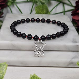 May include: Black onyx bead bracelet with a silver pendant featuring the sigil of the demon Baphomet. The bracelet is on a white marble surface.