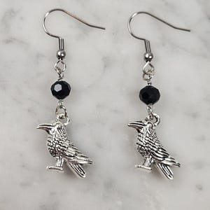 May include: A pair of silver-toned raven earrings. Each earring features a detailed raven charm suspended below a black faceted bead and a silver-colored ear wire. The earrings are designed for pierced ears.