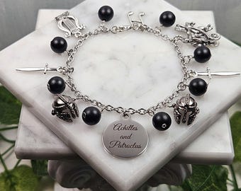 May include: A silver charm bracelet with black beads and various charms, including a lyre, a sword, two helmets, and a round charm with the inscription "Achilles and Patroclus".