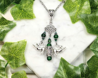May include: Silver-toned pendant necklace with a decorative design. The necklace features green beads and a central cylindrical charm with a symbol. Two cat charms are suspended from the pendant. The necklace is displayed on a white surface with green leaves.
