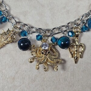 May include: A silver-toned chain bracelet featuring various charms. Gold-colored charms include a seashell, octopus, and trident. Blue beads and crystals are interspersed throughout the design, creating a nautical theme.