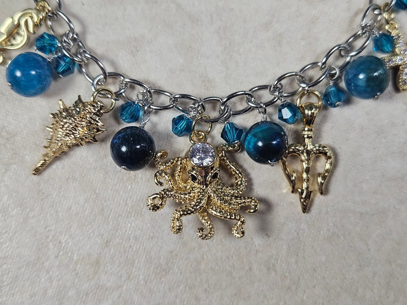 May include: A silver-toned chain bracelet featuring various charms. Gold-colored charms include a seashell, octopus, and trident. Blue beads and crystals are interspersed throughout the design, creating a nautical theme.