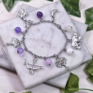 Può includere: Un braccialetto con ciondoli in argento con pietre viola e bianche, con ciondoli a forma di cervo, luna crescente, albero, fungo, orso e freccia e arco.