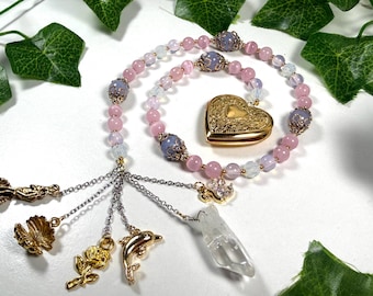 May include: A pink beaded necklace with a gold heart-shaped locket and various gold charms, including a rose, dolphin, and seashell. The necklace also features lavender and clear beads, and a clear crystal pendant.