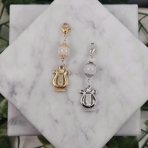 May include: Two silver and gold charm necklaces with a clear crystal bead and a harp charm. The necklaces are on a white marble surface.