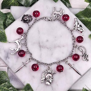 Puede incluir: Un brazalete de plata con cuentas de vidrio rojas y varios dijes de plata, incluyendo un lobo, una serpiente, una espada y un casco.