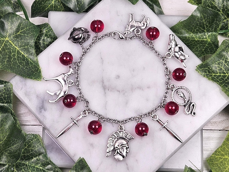 Puede incluir: Un brazalete de plata con cuentas de vidrio rojas y varios dijes de plata, incluyendo un lobo, una serpiente, una espada y un casco.