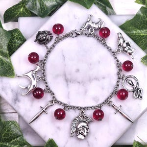 Puede incluir: Un brazalete de plata con cuentas de vidrio rojas y varios dijes de plata, incluyendo una espada, una serpiente, un perro, un casco y un escarabajo.