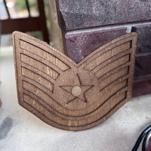 Peut inclure: Insigne de grade militaire en bois, avec une étoile et des rayures. Le bois en couches est teinté brun foncé. Il s'agit probablement d'un objet décoratif ou d'une œuvre d'art sur le thème militaire.