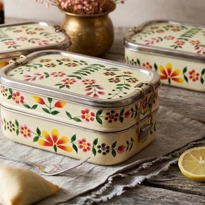 May include: Three rectangular, two-tiered metal lunch boxes with rounded corners, featuring a cream-colored base and a hand-painted floral pattern in red, yellow, and green. Each container has a silver-toned metal rim and secure side latches.
