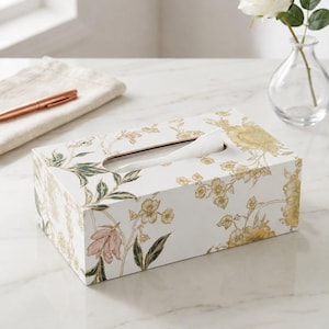 May include: A rectangular tissue box cover featuring a white base with a botanical print of yellow and pink flowers, green leaves, and thin stems. The box has a central oval opening at the top with a single white tissue partially pulled through.