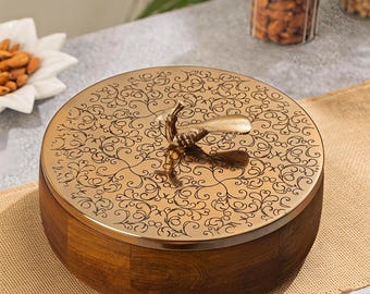 Puede incluir: Un recipiente de almacenamiento redondo de madera con una tapa de metal pulido en tono dorado. La tapa presenta un intrincado patrón de filigrana negra y un asa decorativa central con forma de pavo real. La base es de madera marrón oscura segmentada.