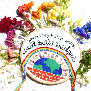 May include: A circular wooden embroidery hoop containing white fabric with hand-stitched text that reads, "when they build walls, we'll build bridges." The design features a colorful rainbow arching over a blue and green globe with small figures.