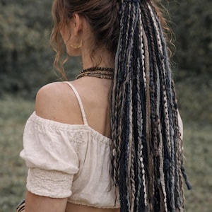 May include: A woman with brown hair in a bun, with long, multi-coloured dreadlocks. The dreadlocks are in shades of blue, brown, and white. She wears a white off-the-shoulder top and gold hoop earrings. The background is a blurred green.