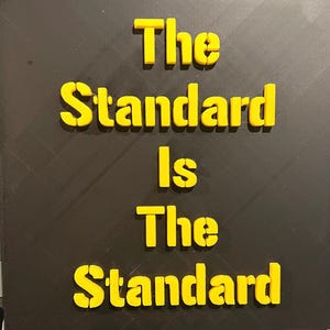 Può includere: Un quadrato nero con il testo giallo "The Standard Is The Standard". Le lettere sono in rilievo e sembrano stampate in 3D. Lo sfondo presenta un sottile motivo strutturato.