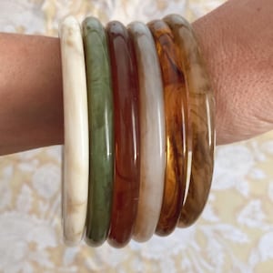 Könnte beinhalten: Eine Sammlung von sechs farbenfrohen Armreifen in Creme-, Grün-, Rot- und Brauntönen. Die Armbänder bestehen aus glänzendem Material und sind an einem Handgelenk gestapelt. Die Armreifen sind etwa 1,3 cm breit.