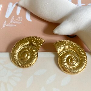 Peut inclure: Une paire de boucles d'oreilles en forme de coquille de couleur or. Les boucles d'oreilles sont détaillées avec un motif en spirale.