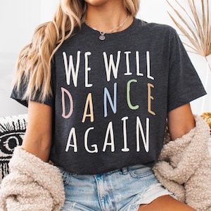 May include: A woman wearing a dark grey t-shirt with the text "WE WILL DANCE AGAIN" in colourful letters. The shirt is a cropped style and the woman is wearing denim shorts.