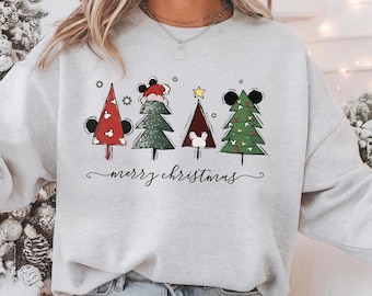 May include: A light gray sweatshirt featuring four Christmas trees with Mickey Mouse ears. The trees are red, green, and brown, with a star and snowflakes. The words "merry christmas" are written in a script font below the trees.