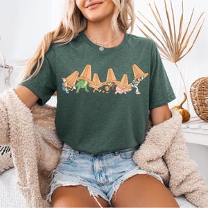 May include: Dark green t-shirt featuring a cartoon graphic of various characters wearing orange traffic cones. The characters are in a line, with a dinosaur, pig, and other figures. The shirt is paired with denim shorts.