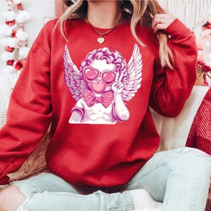 a woman sitting on a couch wearing a red sweatshirt with an angel on it